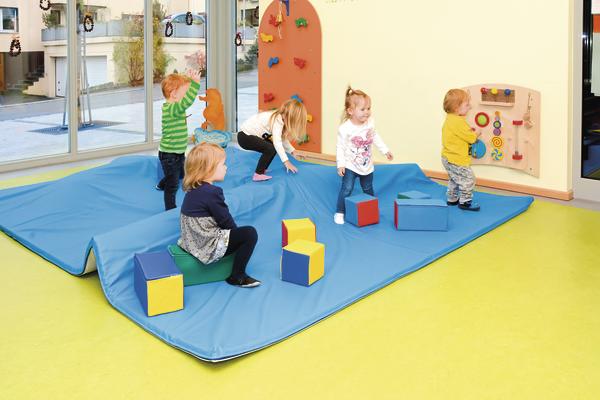 Kinder spielen in einer hellen Kita auf weichen Matten mit bunten Spielwürfeln und Kletterwand.