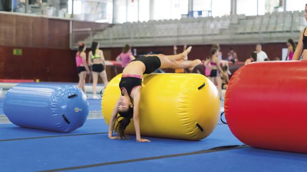 Kind macht Handstand auf großem gelben Gymnastikball in Turnhalle mit anderen Kindern.