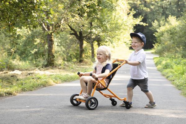 Kind schiebt lachendes Mädchen im TopTrike Buggy auf sonnigem Waldweg, spielerisch und fröhlich.