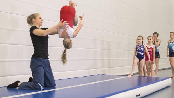Trainer unterstützt Mädchen bei Rückwärtssalto auf Gymnastikmatte, andere Kinder beobachten.