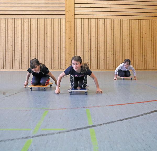 Drei Kinder fahren auf Rollbrettern in einer Turnhalle mit Holzverkleidung.