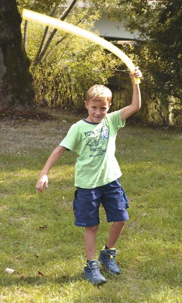 Junge spielt draußen mit gelbem Schaumstoffschwert auf grünem Rasen im Sonnenschein.