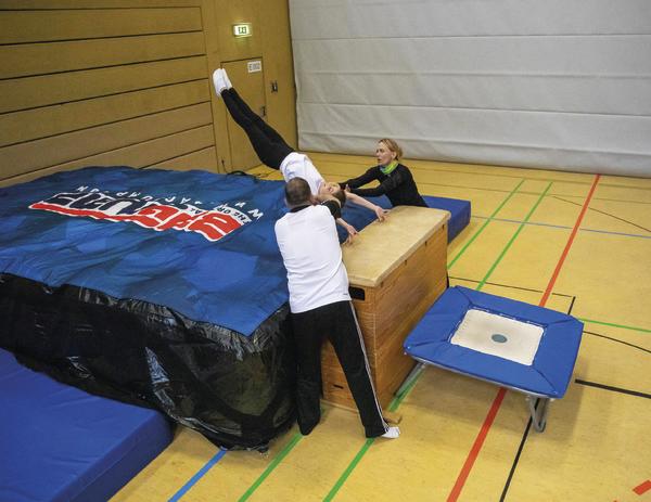 Turnerin macht Rückwärtssalto mit Hilfe von zwei Trainern in einer Sporthalle.
