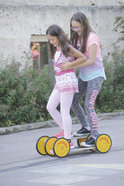 Zwei Mädchen fahren gemeinsam auf einem gelben Pedalo-Spielgerät auf der Straße.
