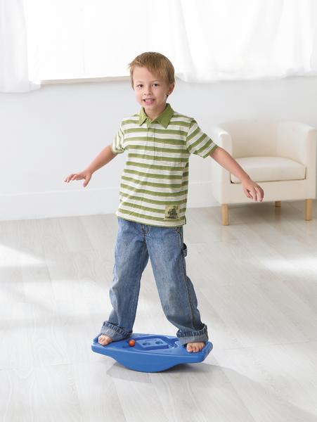 Junge balanciert auf blauem Wackelbrett in hellem Wohnzimmer mit weißem Sessel.