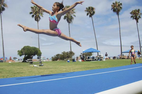 Mädchen springt im Spagat auf blauer Gymnastikmatte im Freien mit Palmen im Hintergrund.
