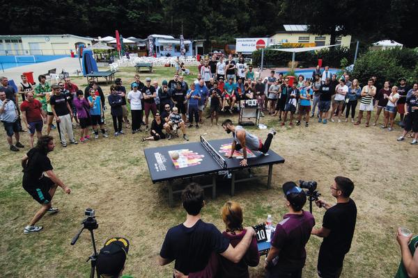 Menschen beobachten ein Outdoor-Tischtennisturnier auf einer Wiese mit Zuschauern und Fotografen.
