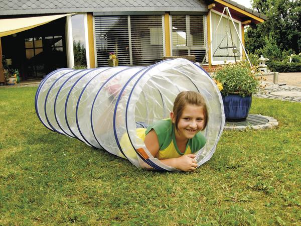 Mädchen krabbelt durch transparenten Spieltunnel im Garten vor Haus mit Terrasse.