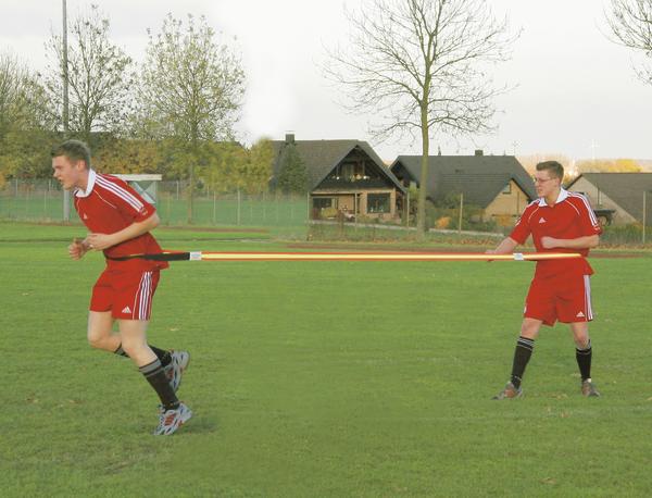 Zwei Fußballspieler in roten Trikots trainieren mit einem elastischen Widerstandsband auf grünem Spielfeld.