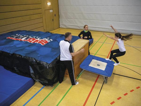 Kind springt auf Trampolin in Turnhalle, betreut von zwei Erwachsenen bei Sportübung.