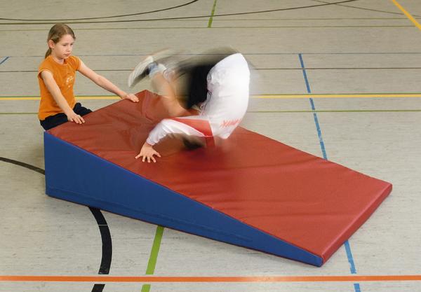 Kind übt Handstand auf roter Schrägmatte in Turnhalle, andere Kind hält Matte fest.