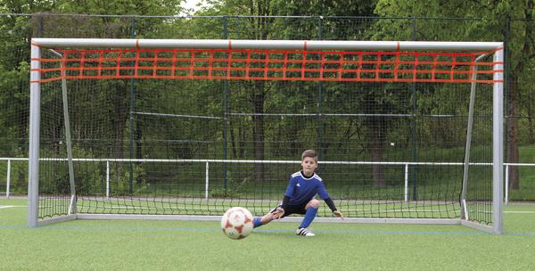 Junger Torwart im blauen Trikot hält Ball vor Fußballtor auf grünem Spielfeld.