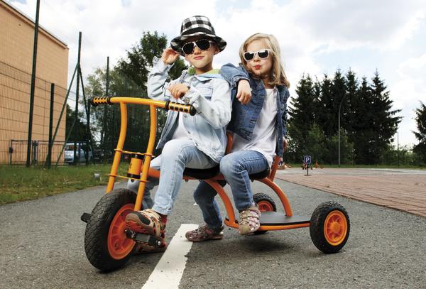 Kinder fahren auf einem orangen Dreirad im Freien, beide tragen Sonnenbrillen und Jeansjacken.