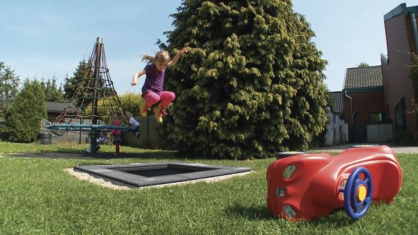 Kind springt auf Trampolin im Garten mit Spielgeräten und rotem Spielauto im Vordergrund.