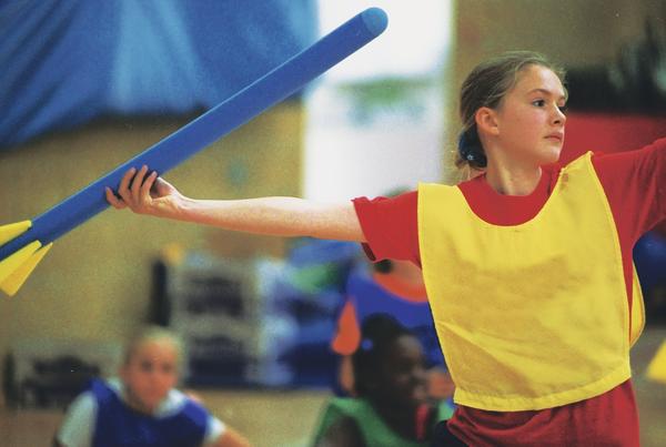 Mädchen in Sportkleidung wirft blauen Schaumstoff-Wurfstab in der Turnhalle beim Sportunterricht.