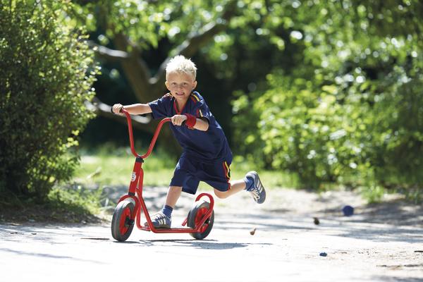 Junge fährt mit rotem Tretroller auf sonnigem Weg im Park, umgeben von grünen Bäumen.