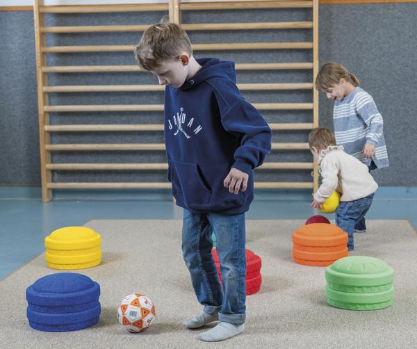 Kinder spielen in einer Turnhalle mit bunten Schaumstoffscheiben und Bällen auf einer Gymnastikmatte.