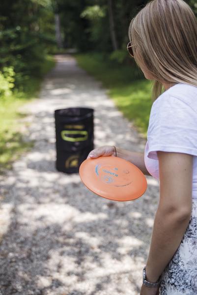 Frau wirft Frisbee auf Waldweg, Ziel ist ein schwarzer Korb für Discgolf.