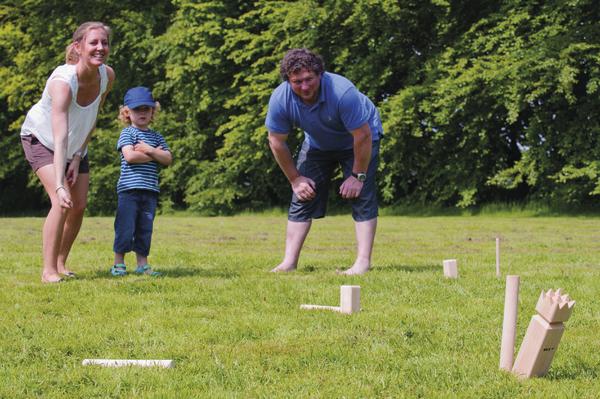 Familie spielt gemeinsam Kubb auf einer Wiese und wirft Holzstäbe auf die Klötze.