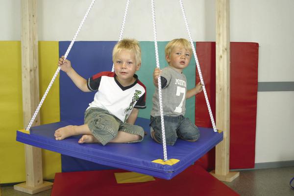 Zwei kleine Jungen sitzen auf einer blauen Therapieschaukel in einem Spielraum.