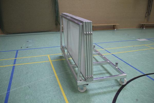 Transportwagen mit zusammengeklappten Tischtennisplatten in einer Sporthalle auf grünem Boden.