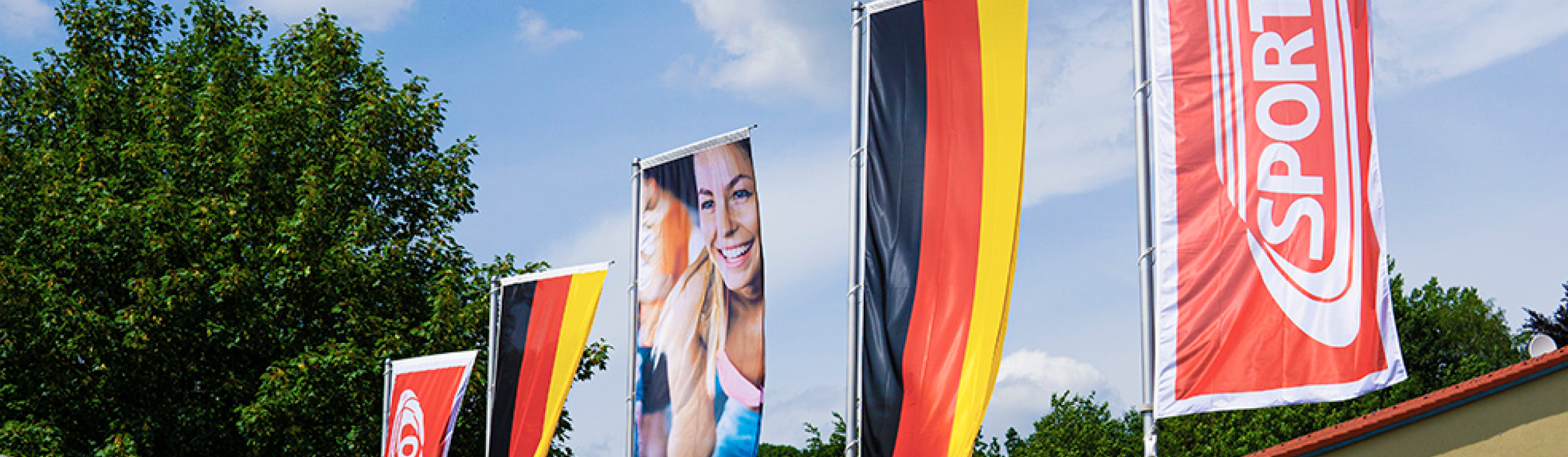 Sportco-Fahnen wehen im WInd, im Hintergrund Bäume und blauer Himmel.