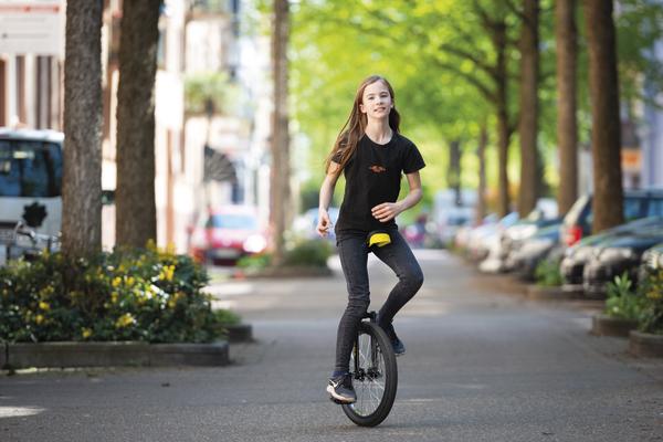 Mädchen fährt Einrad auf städtischem Gehweg, umgeben von Bäumen und parkenden Autos.