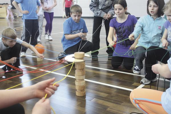 Kinder spielen kooperatives Geschicklichkeitsspiel mit Holzklötzen und Seilen in Turnhalle.