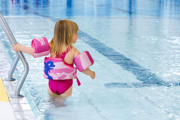 Kleines Mädchen mit Schwimmhilfe und Schwimmflügeln im Schwimmbad am Beckenrand.