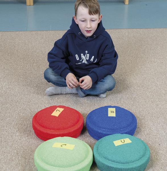 Junge sitzt in einer Turnhalle vor vier farbigen Schaumstoffscheiben mit Buchstabenaufklebern.