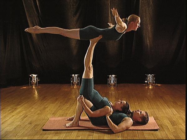 Drei Personen führen gemeinsam eine Akrobatik-Yoga-Pose auf einer Matte in einem Studio aus.