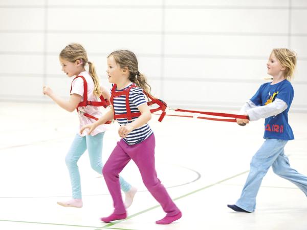 Kinder spielen in der Turnhalle mit Laufgeschirr und roten Bändern beim Bewegungstraining.
