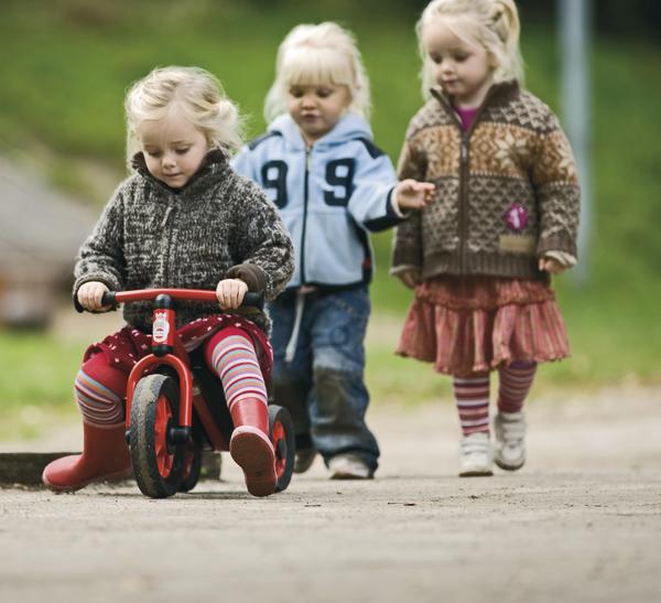 Drei Kleinkinder spielen draußen, eines fährt Laufrad, zwei andere laufen auf Weg.