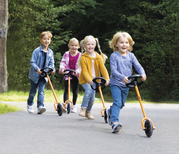 Vier Kinder fahren mit Tretrollern auf einem Weg im Park, umgeben von Bäumen.