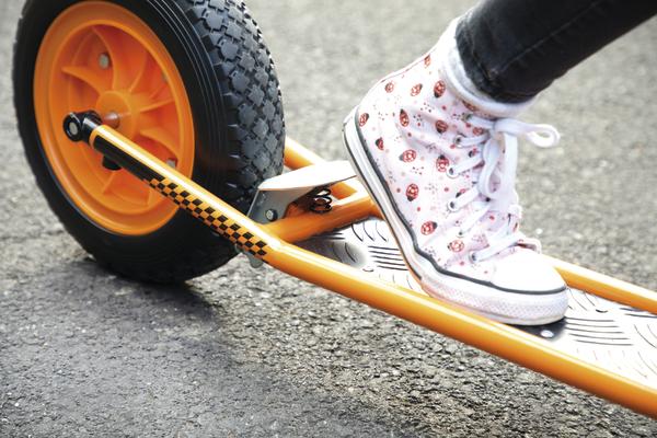 Kind mit weißen Sneakers fährt auf orangefarbenem Tretroller mit großem Reifen auf Asphalt.
