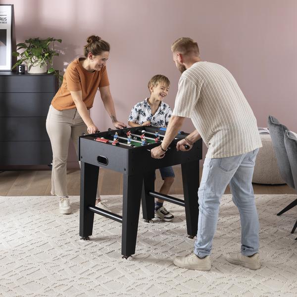 Familie spielt Tischfußball an einem schwarzen Kickertisch im Wohnzimmer.