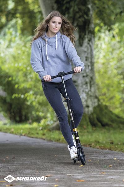 Junge Frau fährt mit schwarzem Kickroller auf Waldweg, trägt grauen Hoodie und Jeans.