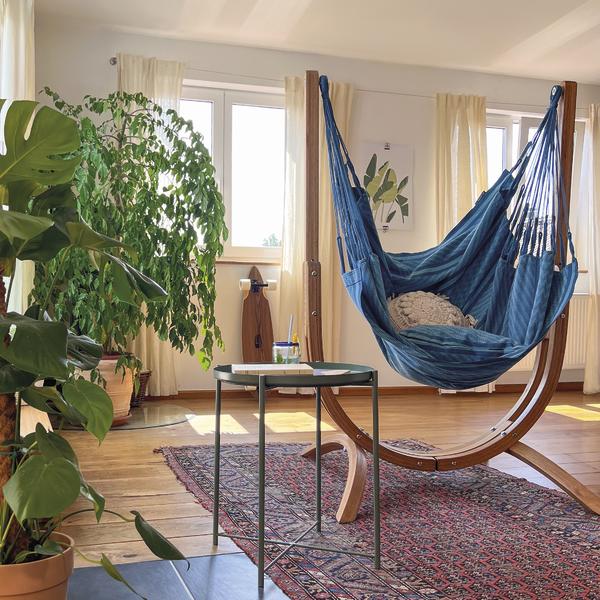 Gemütliches Wohnzimmer mit blauer Hängematte, Pflanzen und Teppich vor großen Fenstern.