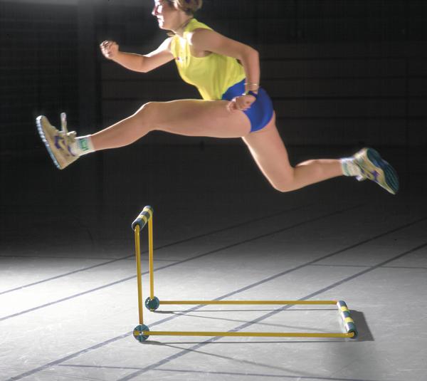 Frau in Sportkleidung springt über Hürde in Halle, dynamische Leichtathletik-Übung.