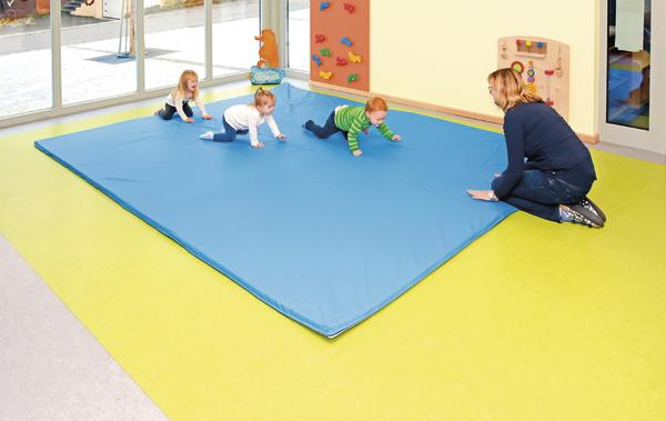 Kinder krabbeln auf blaue Matte im hellen Spielraum, betreut von Erzieherin.