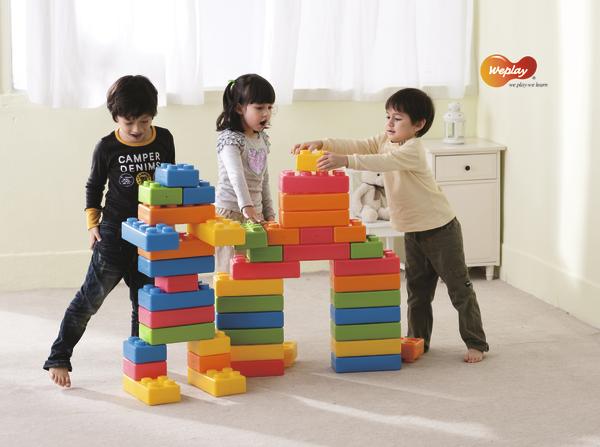 Drei Kinder bauen mit bunten großen Bausteinen eine bunte Brücke im Wohnzimmer.