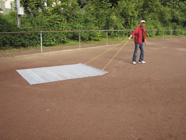 Mann zieht Metallgitter über roten Sportplatz zur Platzpflege bei sonnigem Wetter.