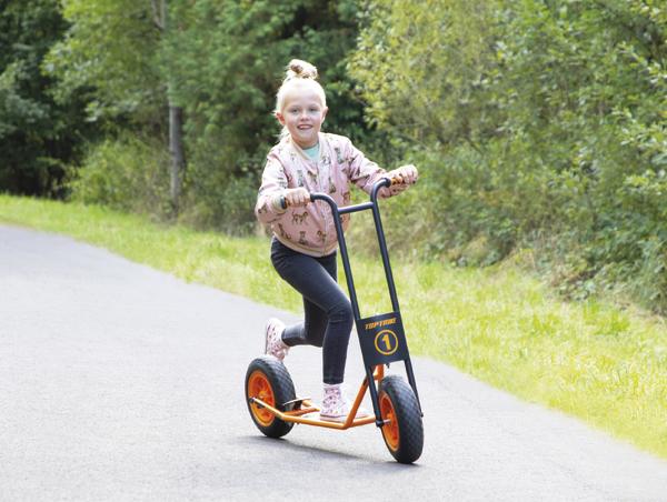 Mädchen fährt auf einem orangefarbenen Tretroller auf einem Waldweg bei Tageslicht.