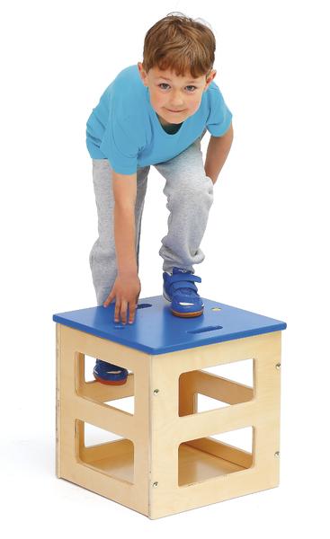 Junge klettert auf bunten Holzspielwürfel, trägt blaues Shirt und graue Hose.