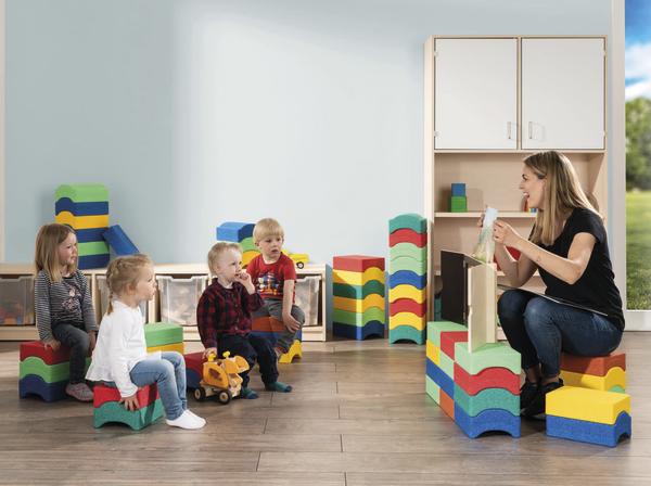 Erzieherin zeigt einer kleinen Kindergruppe auf bunten Schaumstoffhockern etwas auf einem Blatt Papier.