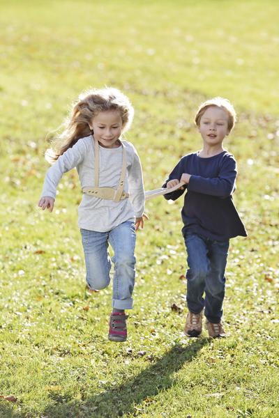 Zwei Kinder laufen lachend über eine sonnige Wiese im Freien.