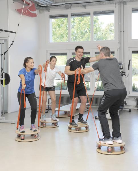 Vier Personen trainieren mit Balance-Boards und Widerstandsbändern in einem hellen Fitnessraum.