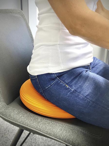 Person sitzt auf einem orangen Sitzkissen auf einem grauen Stuhl zur Haltungskorrektur.
