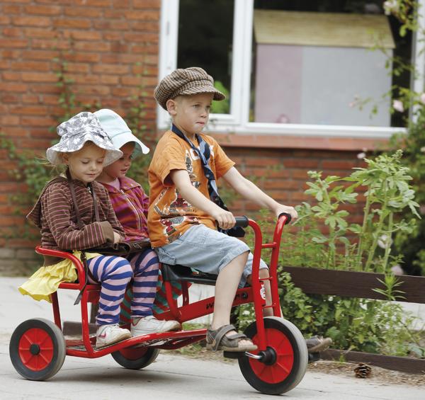 Drei Kinder fahren gemeinsam auf einem roten Dreirad vor einem Backsteinhaus.