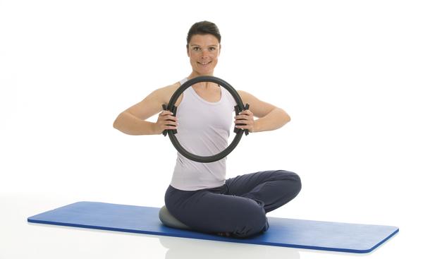 Frau macht Pilates-Übung mit Pilates-Ring auf blauer Matte, weiße Sportkleidung.